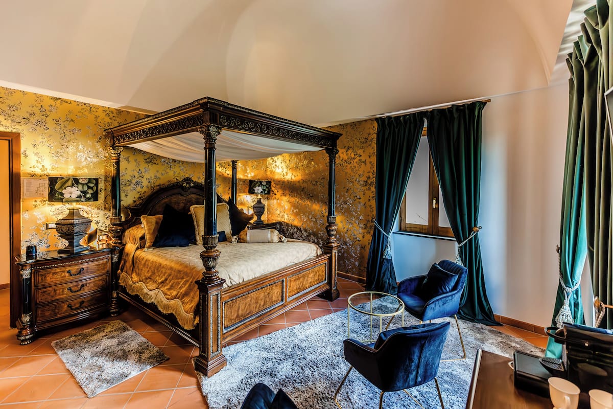Un lit à baldaquin, un fauteuil bleu foncé, une petite table ronde et un ordinateur portable posé sur une table de chevet en bois.