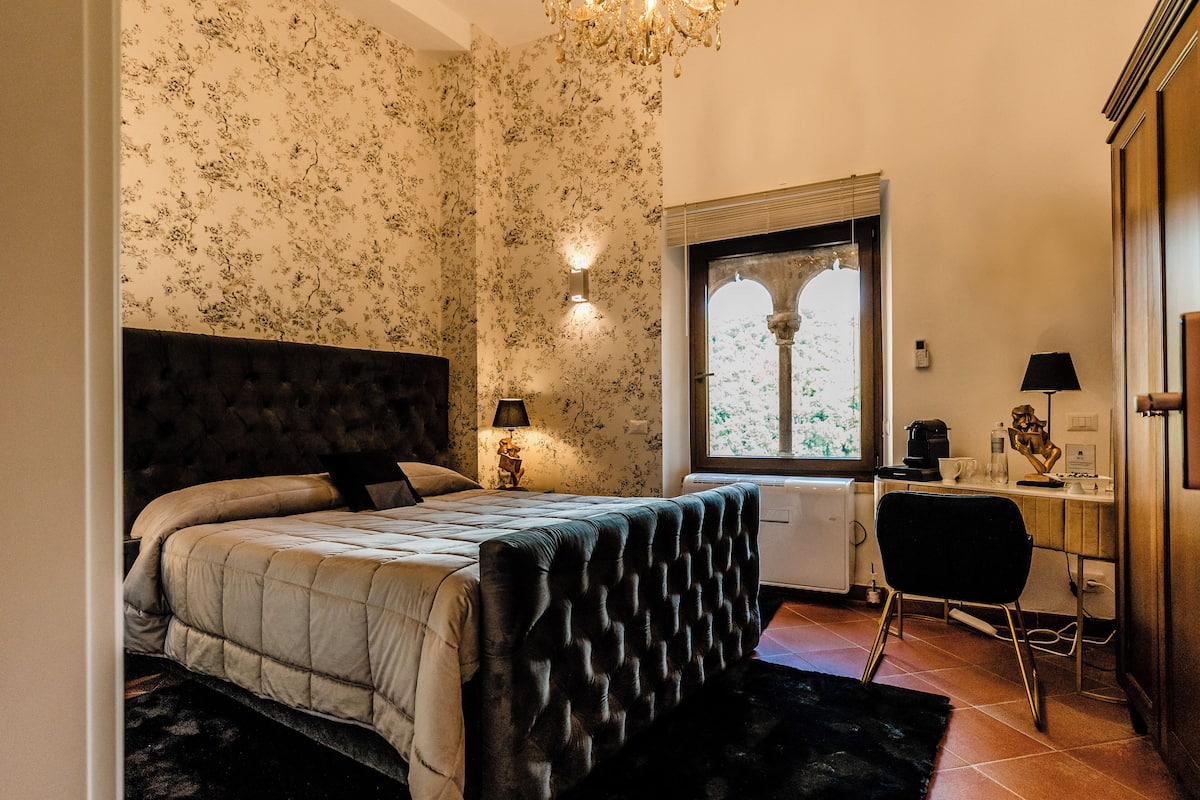 Une chambre avec un grand lit, un bureau et une chaise. Il y a une fenêtre donnant sur un paysage verdoyant.