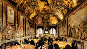 Een statige balzaal met versierde plafondfresken, meerdere lange tafels gedekt voor een banket, en klassieke kunstwerken aan de wanden.