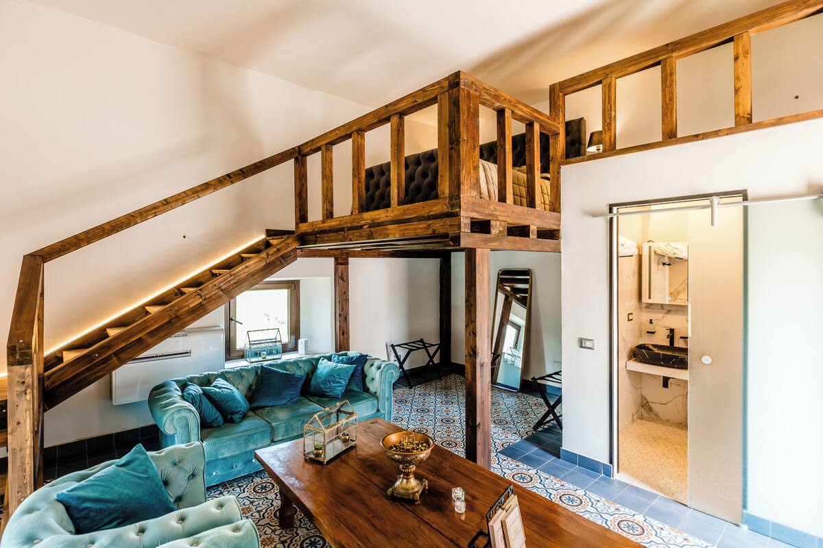 Un vaste espace de vie agrémenté d’un grand escalier en bois, d’un ensemble de canapés bleus, d’une table basse en bois et d’un coin cuisine avec un évier et des placards.