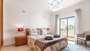 Et soveværelse med en stor seng, to sengeborde med lamper, en stol, en balkon med udsigt og en moderne loftlampe.