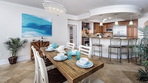 A dining area with a wooden table, white chairs, and blue glassware. There is a kitchen in the background with wooden cabinets and stainless steel appliances. - BR 108: Golden Horizon - Indulge in the Ultimate Beachfront Escape! 2nd level (Fort Walton Beach)