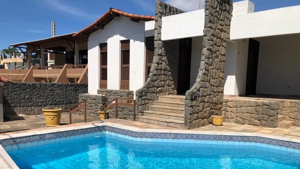 A swimming pool with stone walls and a tiled edge, surrounded by a patio area with steps leading to a building with a white facade and brown shutters. - conceitual hostel natal (Natal)