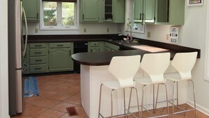 Una cocina con armarios verdes, una isla central con taburetes blancos, un refrigerador y un tragaluz.
