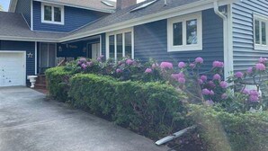 Una casa de dos pisos con garaje, un porche cubierto y un jardín bien cuidado con flores moradas.