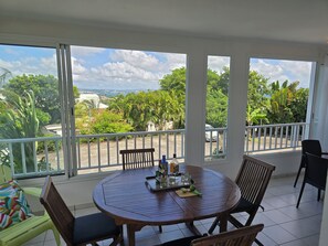 A round wooden table with chairs, a balcony with a railing, and a view of greenery and buildings.