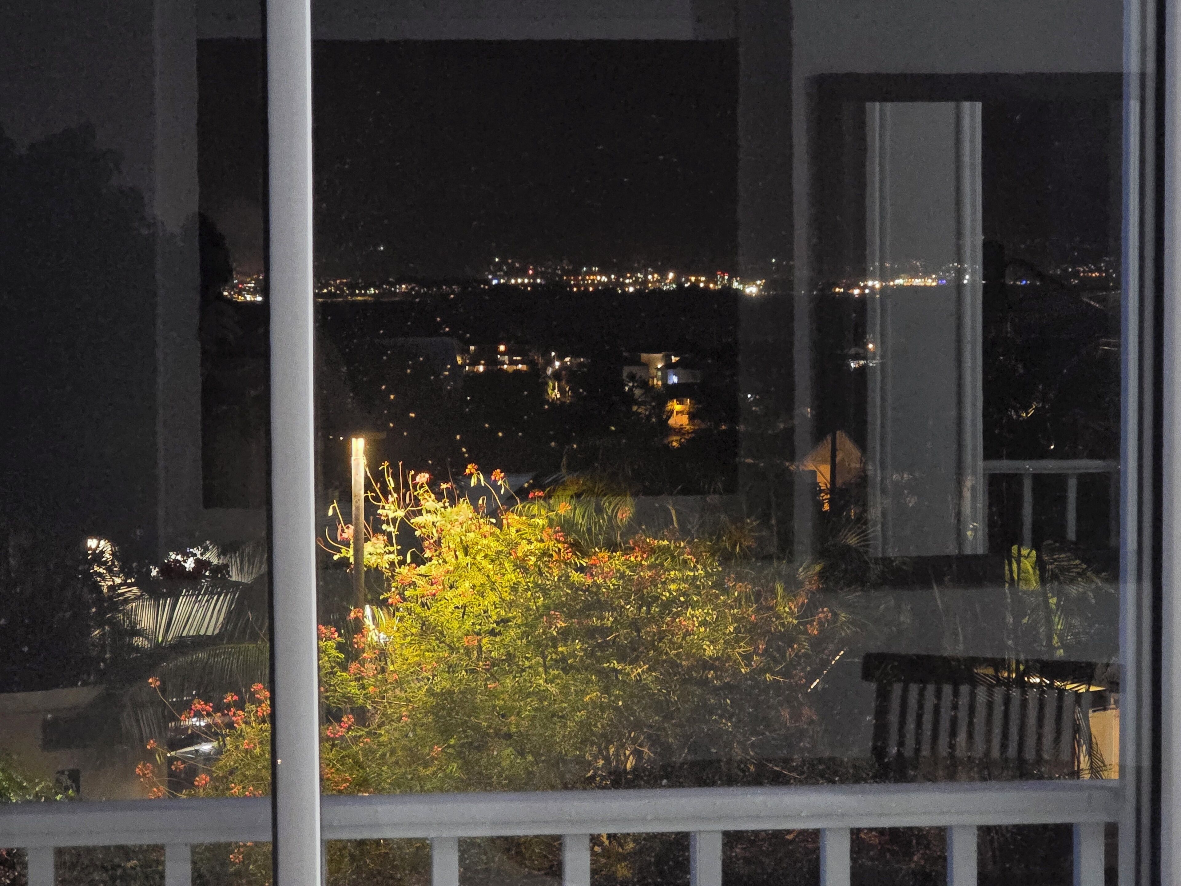 Vista nocturna a través d'una finestra amb un balcó, un paisatge urbà al fons i vegetació al primer pla.