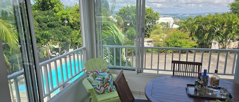 Un balcón con vista a una piscina, una mesa redonda con sillas y una ventana que da a zonas verdes y edificios.