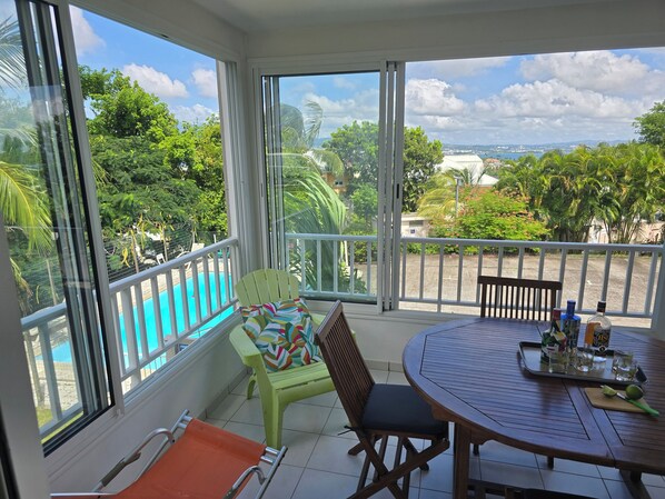 A balcony with a view of a swimming pool, a round table with chairs, and a window with a view of greenery and buildings.