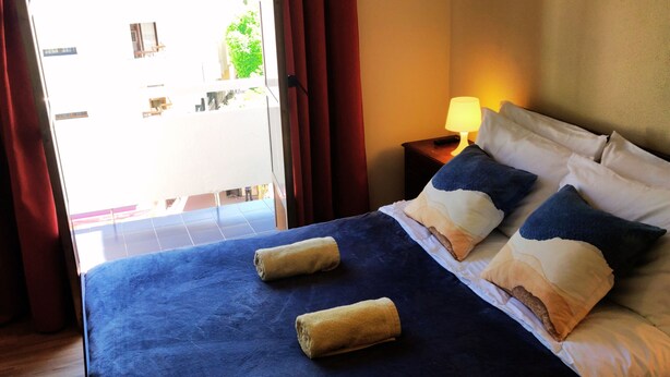 Habitación de hotel con una cama grande, dos toallas, una mesita de noche con lámpara y vistas al balcón exterior.