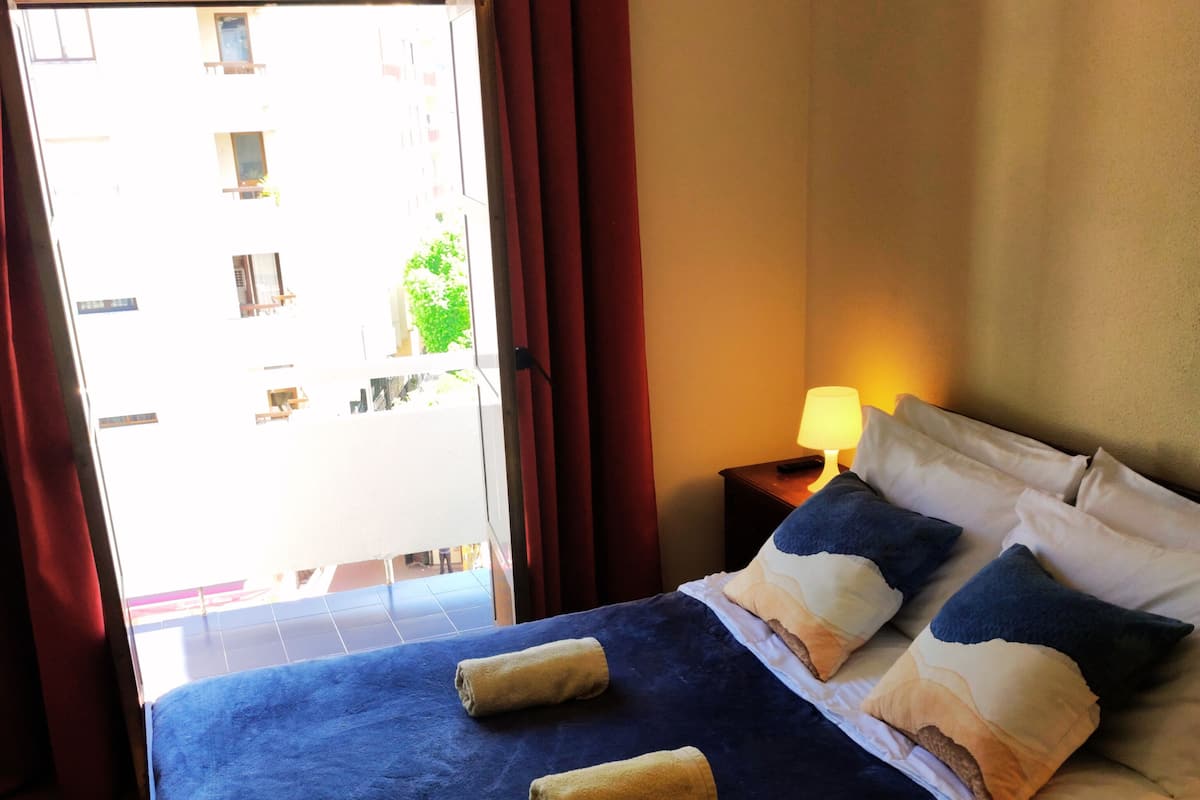 A hotel room with a large bed, two towels, a nightstand with a lamp, and a view of the outside balcony.