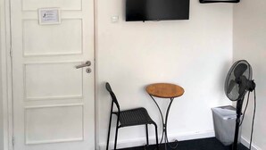 Una habitación con una puerta blanca, una silla negra, una mesita redonda pequeña, un ventilador negro y un televisor montado.