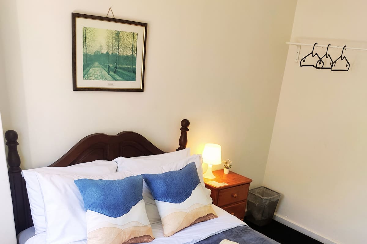 A neatly made bed with a blue blanket, two pillows, and two rolled towels. There is a framed picture on the wall above the bed, a bedside lamp, and a small trash bin.