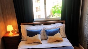 Una cama bien tendida con ropa de cama azul y dos toallas enrolladas, una lámpara de noche y una ventana con cortinas.
