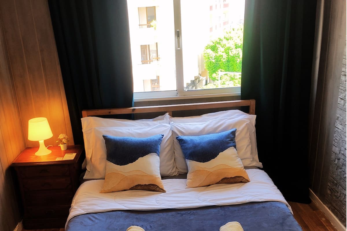 A neatly made bed with blue bedding and two rolled towels, a bedside lamp, and a window with curtains.