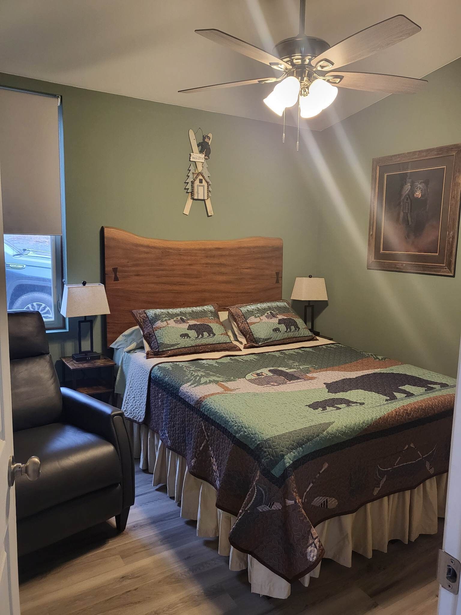 A bedroom with a wooden headboard, a quilted bedspread with animal prints, a ceiling fan, and a welcome sign.