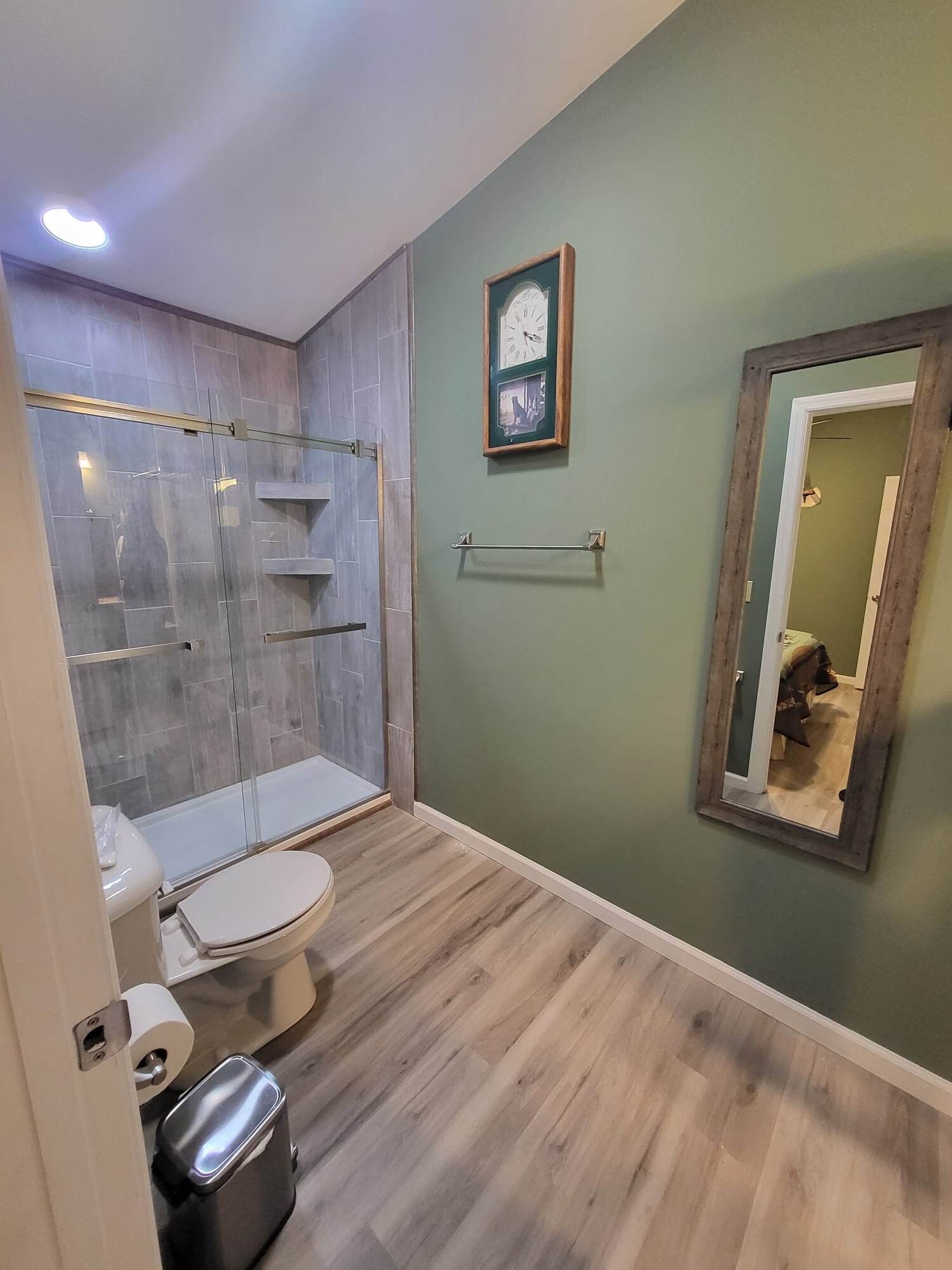 Et badeværelse med glasbrusekabine, toilet, skraldespand og spejl.