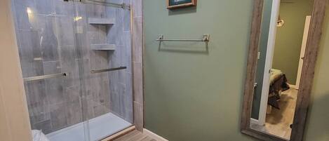 Et badeværelse med glasbrusekabine, toilet, skraldespand og spejl.