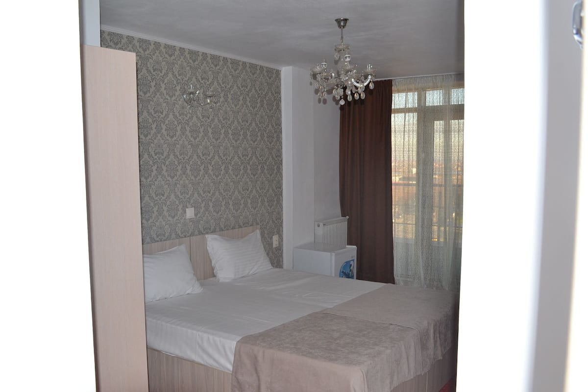 Un dormitorio con cama, radiador, mesita de noche, candelabro y ventana con cortinas.