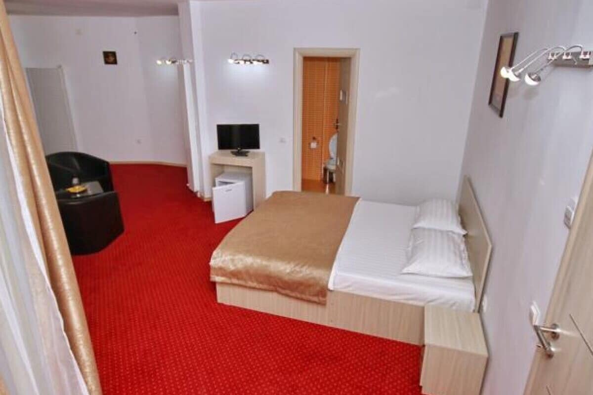 Habitación de hotel con alfombra roja, cama con ropa blanca, mesita de noche pequeña, televisor y armario.