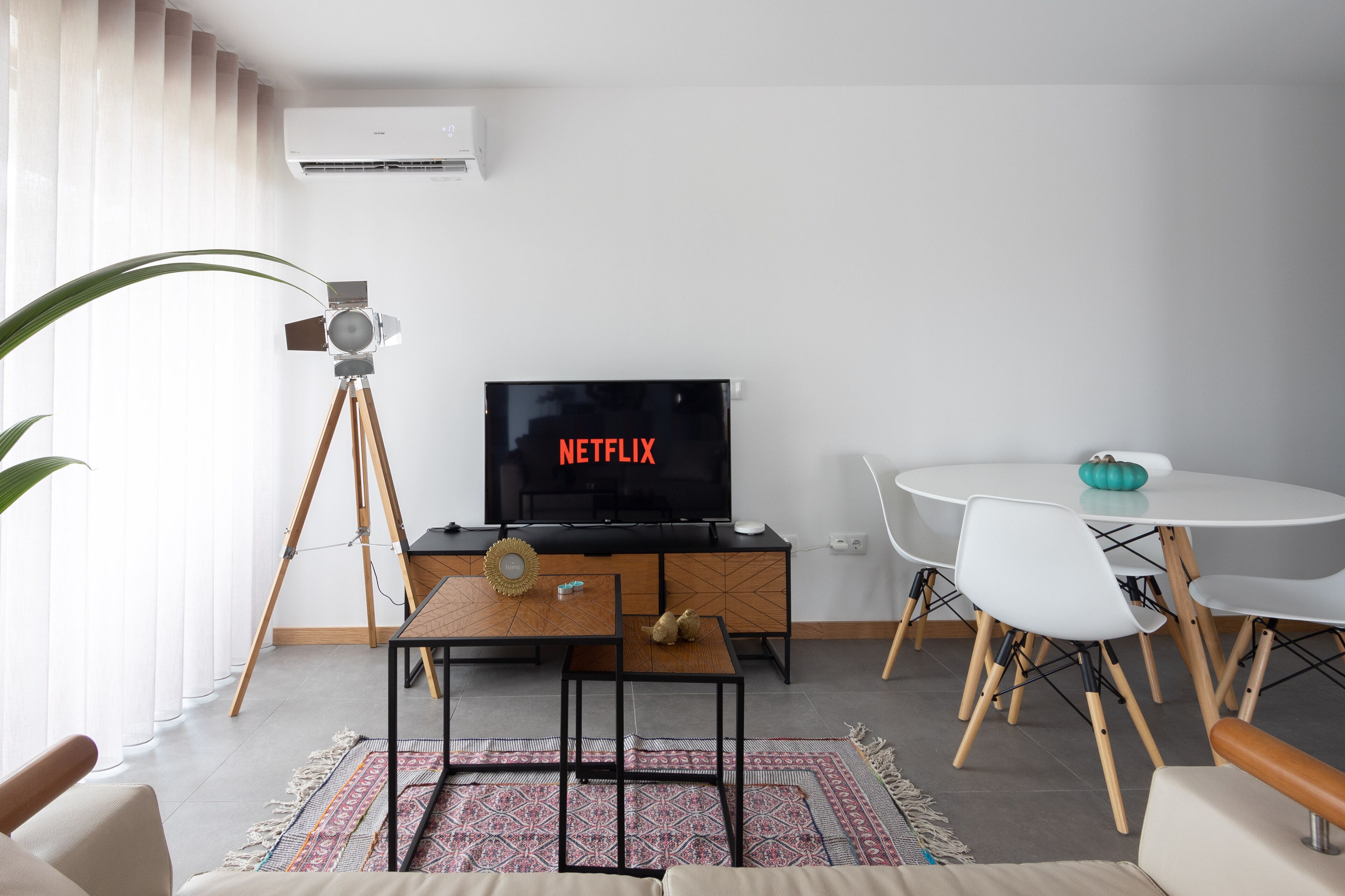 Ett modernt vardagsrum med en platt-TV som visar "NETFLIX", ett soffbord i trä, ett matbord med stolar och en stor krukväxt.