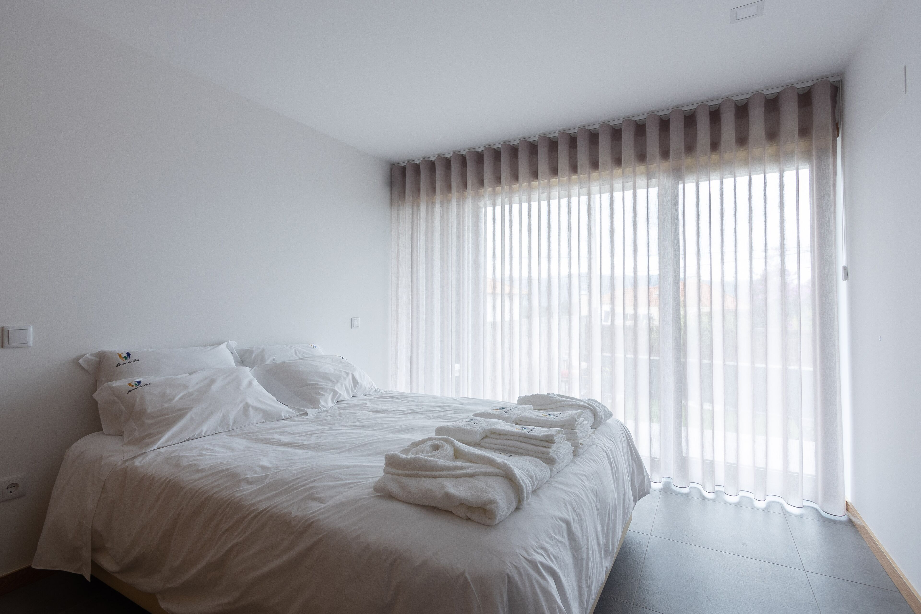 Ein ordentlich bezogenes Bett mit weißen Bettwäsche und Handtüchern, ein Fenster mit durchsichtigen Vorhängen und eine schlichte weiße Wand.