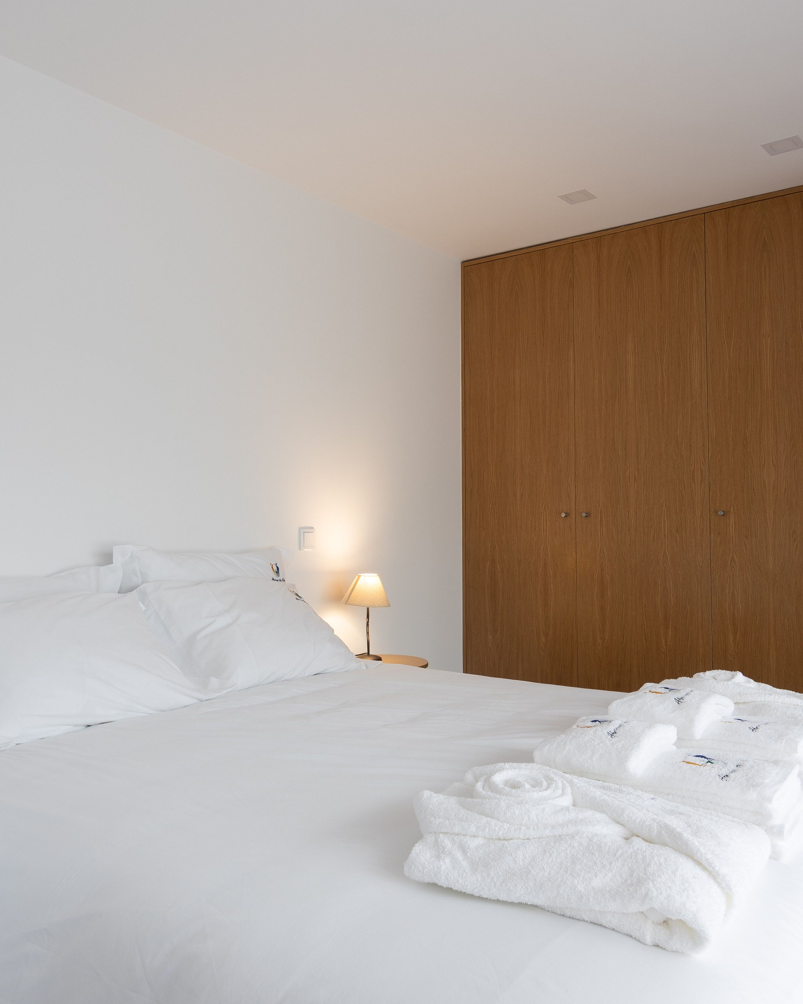 A neatly made bed with white linens and pillows, a wooden wardrobe, and a bedside lamp.