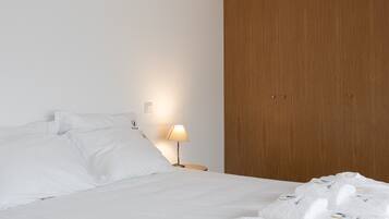 A neatly made bed with white linens and pillows, a wooden wardrobe, and a bedside lamp.