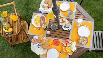 Una mesa de desayuno preparada con platos, pan, frutas y bebidas sobre una mesa de madera al aire libre.
