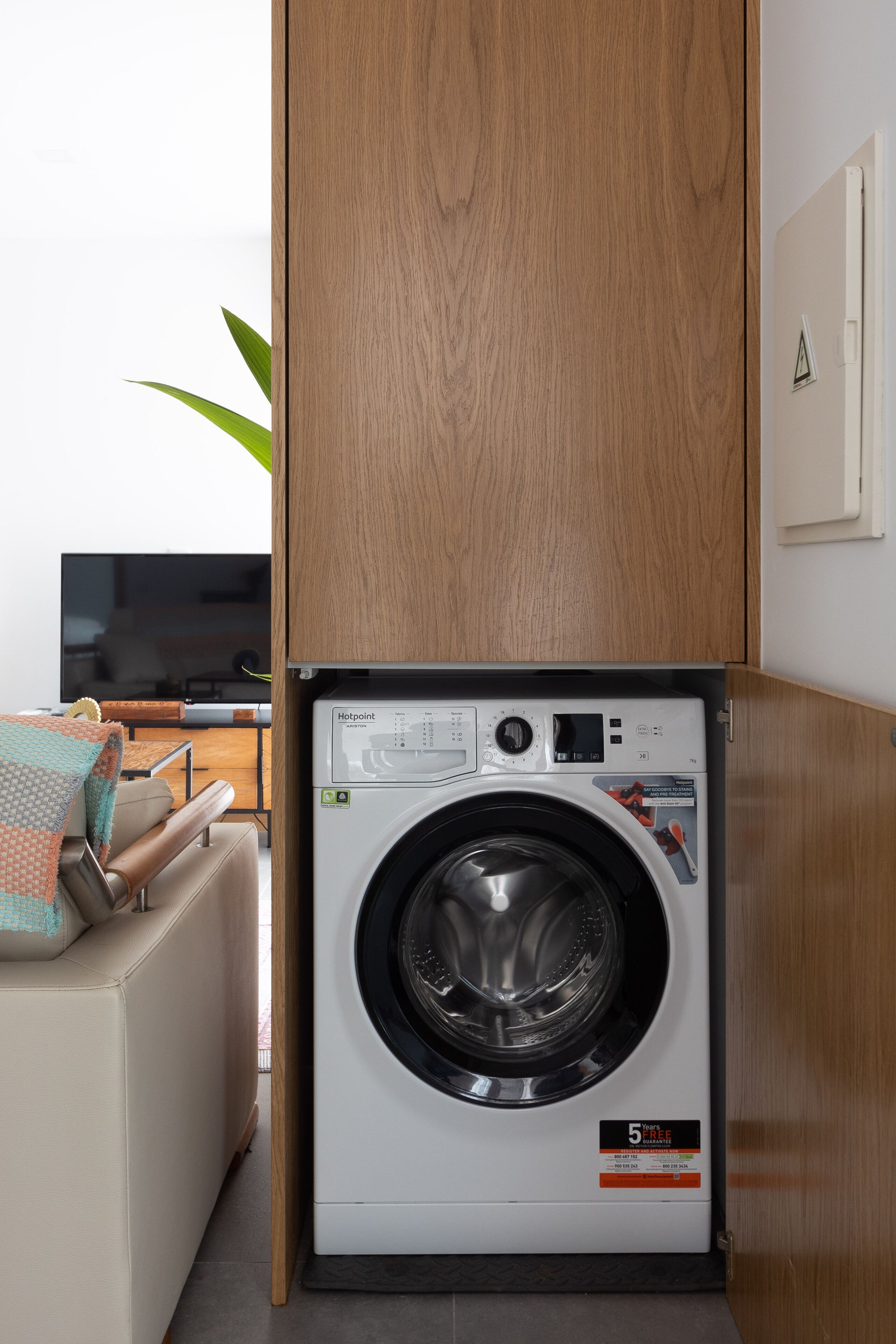 A compact, modern laundry room with a front-loading washing machine, wooden cabinetry, and a seating area with a sofa and a television.