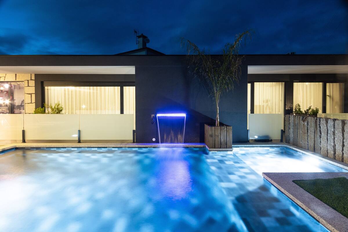 A modern outdoor pool area with a blue-lit water feature, stone walls, and potted plants.