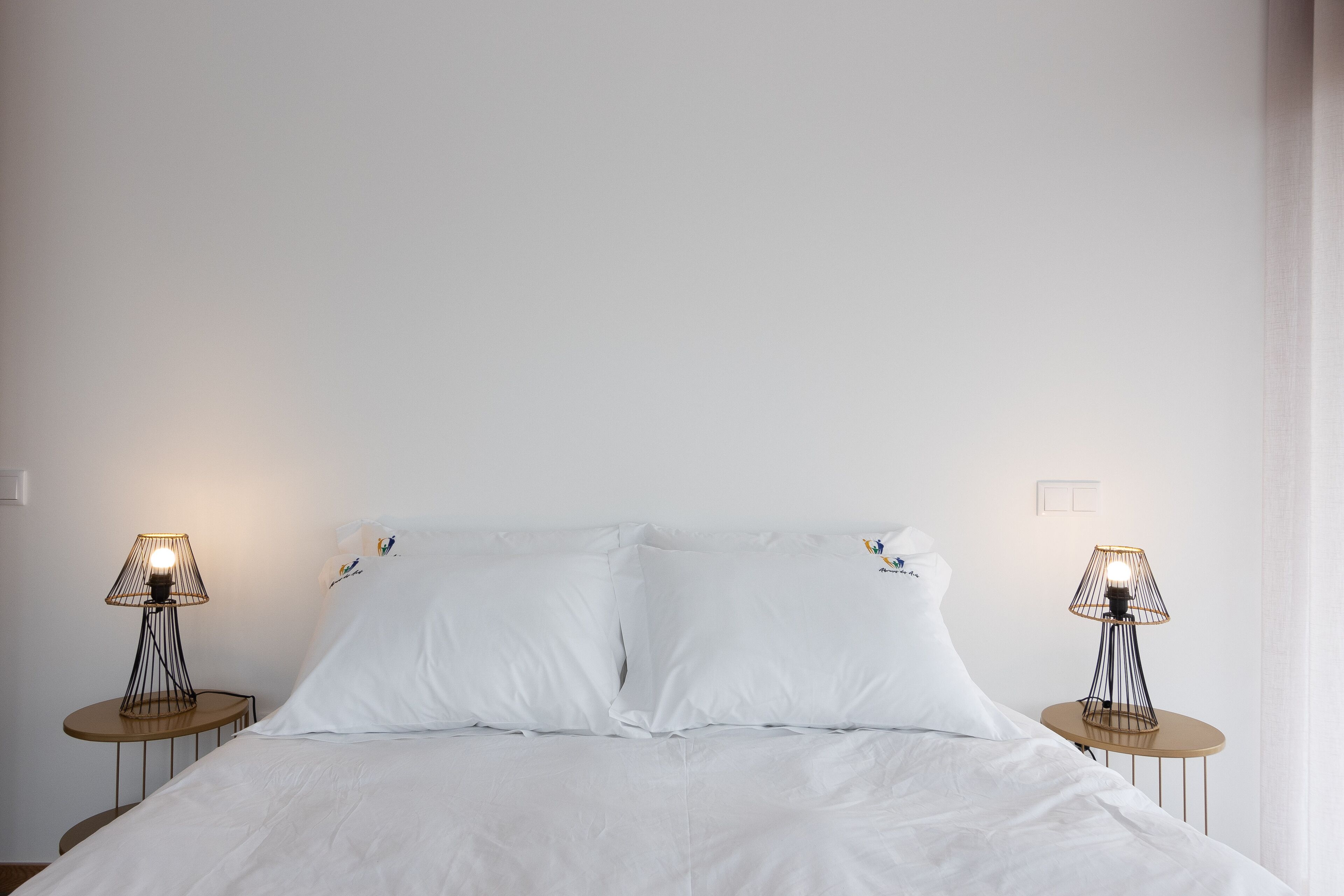 A neatly made bed with white linens, flanked by two bedside tables with lamps, in a simple room with plain walls and minimal decor.
