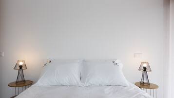 A neatly made bed with white linens, flanked by two bedside tables with lamps, in a simple room with plain walls and minimal decor.