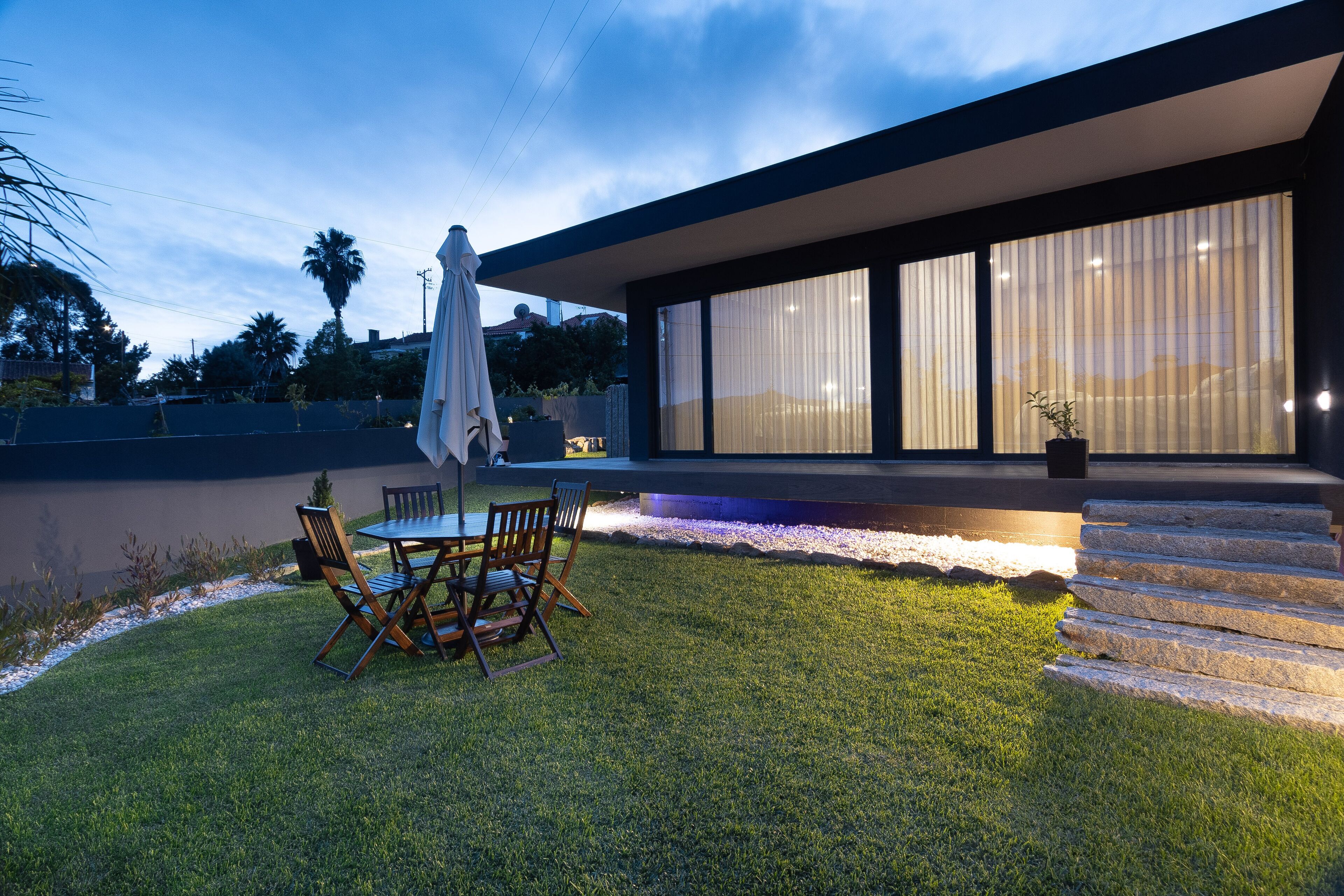 Uma casa moderna com varanda, mesa de jantar com cadeiras e uma grande janela com vista para uma área ajardinada.