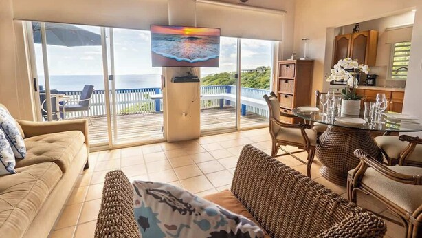 Een ruime woonkamer met een grote televisie, comfortabele zitplaatsen, een eettafel en uitzicht op de oceaan door grote schuifdeuren.