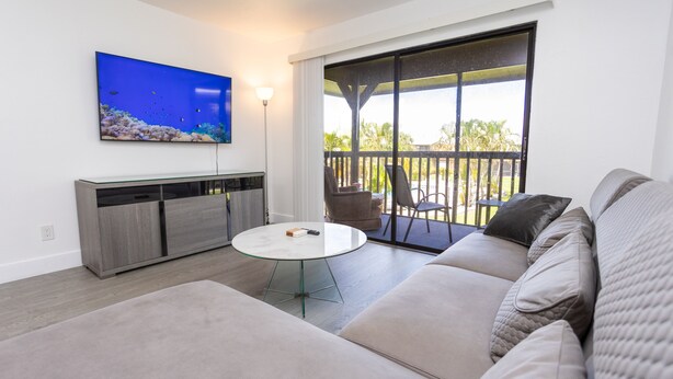 Una sala moderna con un televisor de pantalla plana grande, una mesa de centro de vidrio, un sofá gris y un balcón con mobiliario exterior.