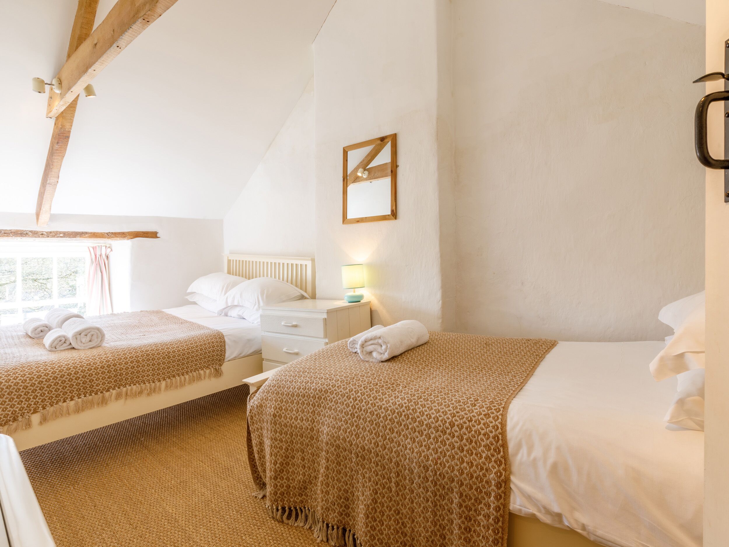 Une chambre avec deux lits, une fenêtre avec des rideaux, une table de chevet avec une lampe et une poutre en bois au plafond.