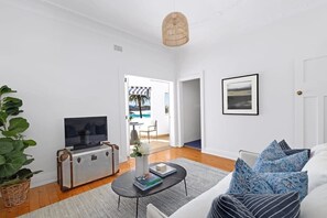 A modern living room with a flat-screen TV on a vintage-style cabinet, a large potted plant, a coffee table with books, and a view of the outdoors through an open door. - Absolute Seafront Bronte (Bronte)