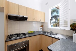 A kitchen with wooden cabinets, a gas stove, a sink, and a window with blinds. - Absolute Seafront Bronte (Bronte)