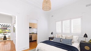 A modern bedroom with a large bed, bedside tables, a window with blinds, and a view of a balcony and kitchen area. - Absolute Seafront Bronte (Bronte)