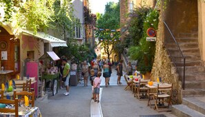A narrow street with outdoor seating, a market stall, and a staircase leading to a building.