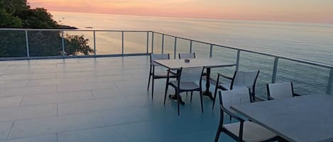 A terrace with a table and chairs overlooking the sea at sunset.
