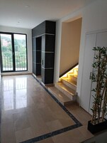 Una habitación amplia con un gran ventanal corredera que da a un balcón, una escalera con escalones iluminados y una planta en maceta.