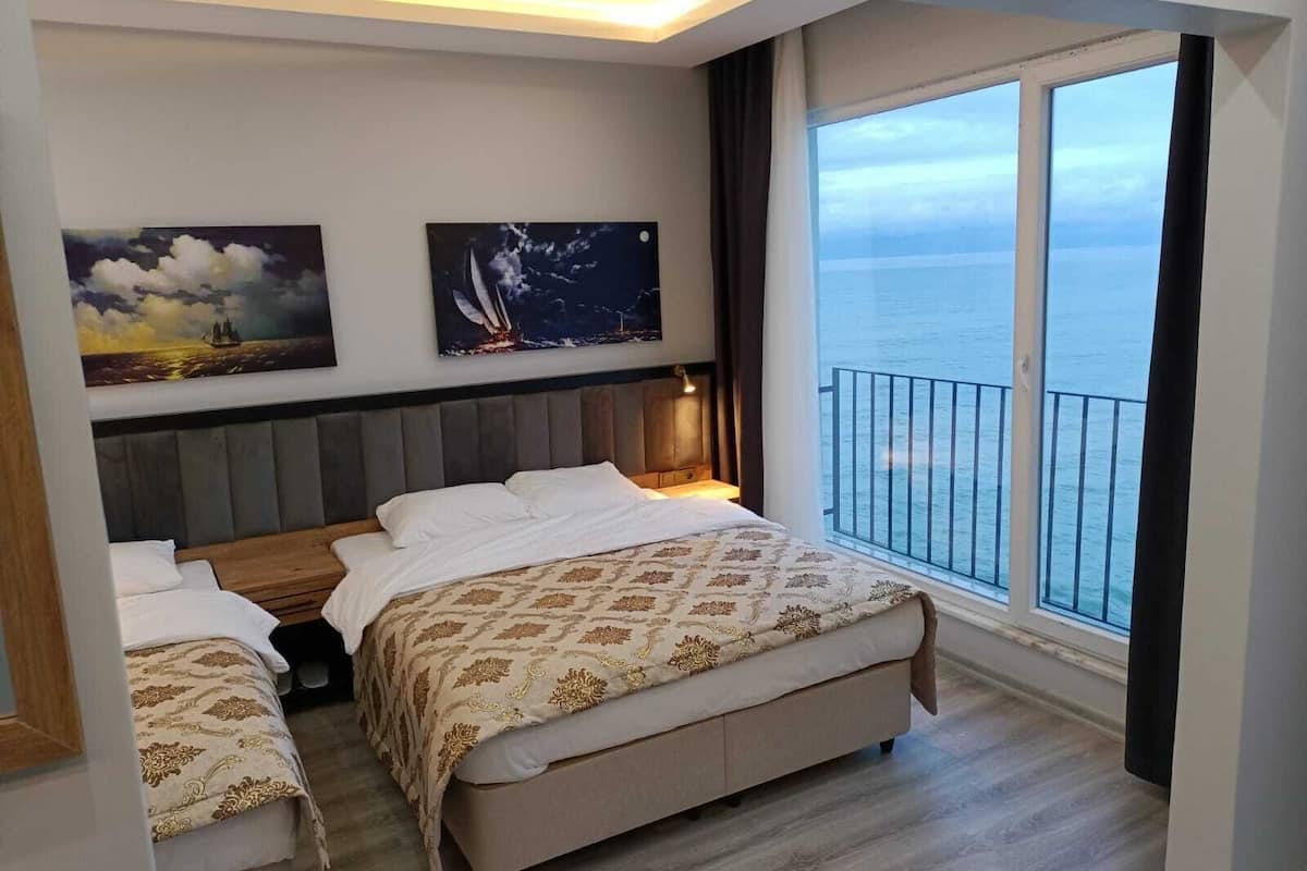 A bedroom with a large window offering a sea view, a bed with a patterned bedspread, and two wall-mounted paintings.