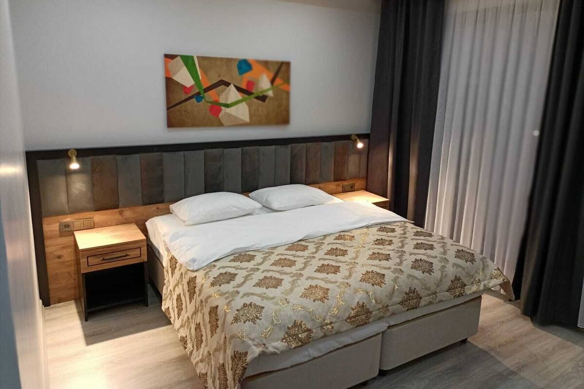 A modern bedroom with a large bed, a nightstand, a headboard with built-in lighting, and a colorful abstract painting on the wall.