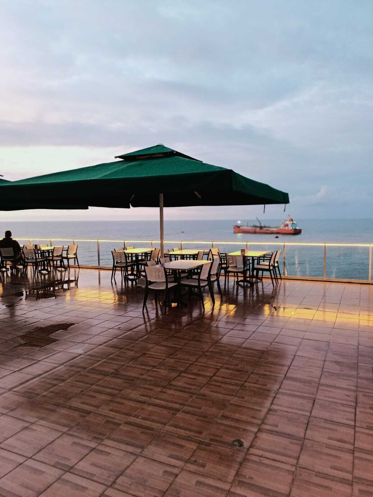 An outdoor dining area with tables and chairs, a green umbrella, and a view of the sea with a ship in the background.