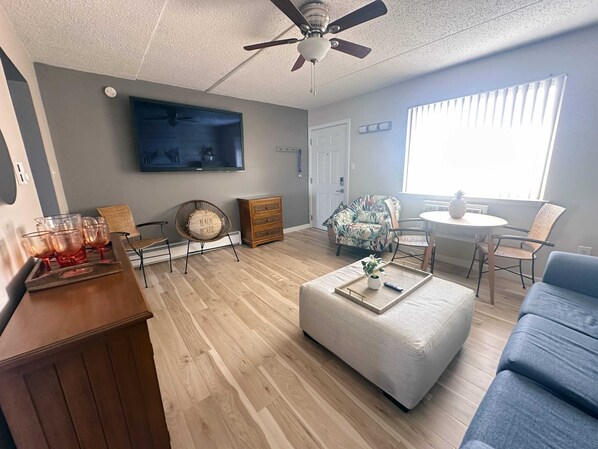 A modern living room with a wooden floor, a ceiling fan, a flat-screen TV, a dining area with a table and chairs, and a large window with blinds. - 1BR Pent. Suite 1 Block Beach/Boards/Fun/Elevator (Ocean City)