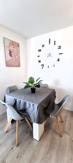 A dining area with a round table covered by a grey tablecloth, two grey chairs, a potted plant, and a large wall clock.