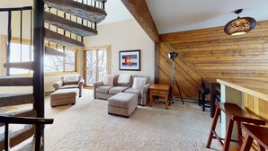 A cozy living room with a staircase, a wooden ceiling, a framed flag, and a floor lamp. - C&C Ski Factory-Ski in/Ski Out (Crested Butte)