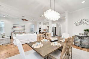 A spacious living room with a large wooden dining table, white sofas, and a chandelier.